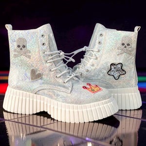 May include: Silver glitter combat boots with white laces and soles. The boots feature decorative patches including a skull and crossbones, a heart, a star, and a crown. The boots have a chunky sole.