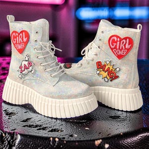 May include: A pair of silver glitter ankle boots with white soles. Each boot features a red heart patch with "GIRL POWER" text and comic-style patches that say "ZAP" and "BANG!"