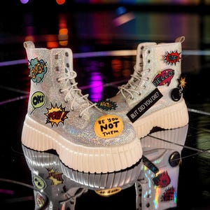 May include: A pair of silver glitter combat boots with white laces and thick, off-white soles. The boots are adorned with comic book-style patches that read "OMG!", "OK!", "POW!", "WIF!", "HELLO", and "BUT DID YOU DIE?" and "BE YOU NOT THEM".