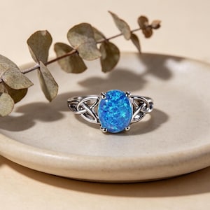 May include: A silver ring with an oval, blue opal stone. The ring features a Celtic knot design on the band. The opal has a speckled appearance, with shades of blue. The ring is displayed on a neutral-colored plate.