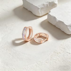 Op de afbeelding: Twee roségouden oorringen met iriserende opaal inlays. De oorbellen worden gepresenteerd op een wit oppervlak met twee grote, witte, onregelmatig gevormde rotsen op de achtergrond. Sieraden.