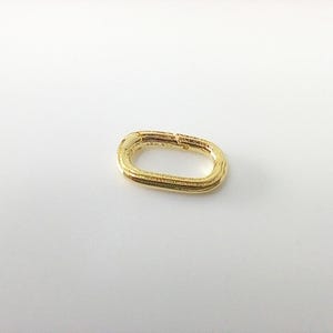 May include: A gold-toned oval-shaped spring ring clasp featuring a hinged opening mechanism. The metal surface has a textured, grooved finish along the outer edge, and the clasp is designed for connecting jewelry components like chains or charms.