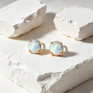 May include: A pair of gold stud earrings with iridescent opal-like stones. The earrings are round with a gold setting and four small gold prongs. The opalescent stones display a range of colors, including blue, pink, and white.