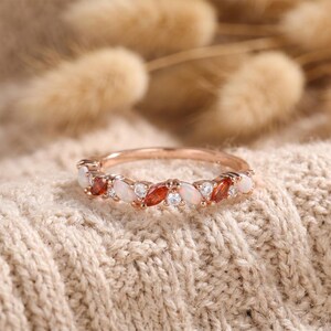May include: Rose gold ring with alternating oval-cut gemstones in white, orange, and clear. The ring is set on a neutral-colored, textured surface. The gemstones are evenly spaced around the band.
