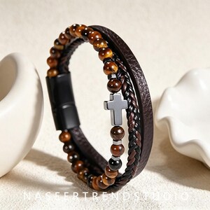 May include: A multi-strand bracelet featuring a row of brown tiger eye beads, a braided dark brown leather cord, and a smooth dark brown leather strap. A small, dark gray metal cross charm is integrated into the beaded strand, secured by a black metal clasp.