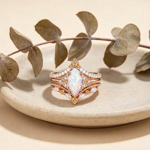 May include: A rose gold ring set featuring an opal center stone and smaller accent stones. The set includes a main ring and two additional bands, all with intricate detailing. The rings are displayed on a neutral-toned plate with dried foliage.