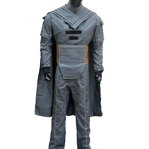 May include: A gray costume with a cape, pants, and a jacket. The jacket has a high collar and a brown belt. The costume is displayed on a black mannequin. White stitching details the seams of the outfit.