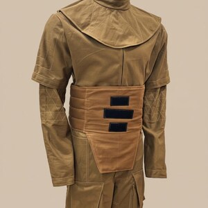 Puede incluir: Un disfraz de color marr&oacute;n con cuello alto, capa en los hombros y cintur&oacute;n ancho con detalles rectangulares negros. La parte superior de manga larga y los pantalones tienen detalles de costura geom&eacute;trica. La cabeza del maniqu&iacute; es dorada.