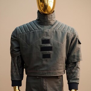 Puede incluir: Maniqu&iacute; con chaqueta y pantal&oacute;n verde oscuro. La chaqueta tiene cuello alto, hombros acolchados y parches negros. Las mangas tienen costuras geom&eacute;tricas. El maniqu&iacute; tiene cabeza y manos doradas.