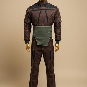 Puede incluir: Mono marr&oacute;n con detalles negros y costuras blancas, exhibido en un maniqu&iacute;. El conjunto incluye una parte superior de cuello alto, un cintur&oacute;n verde ancho y pantalones a juego. El maniqu&iacute; tiene cabeza y manos doradas.