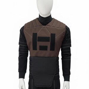 May include: Brown and black tactical vest with a geometric design on a mannequin. The vest has a padded brown front with black rectangular accents and a black lower section. The mannequin is wearing a black long-sleeved shirt and pants.