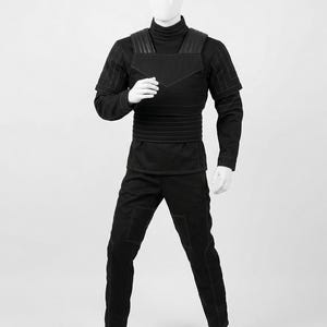 Pode incluir: Um traje preto exibido em um manequim. O traje inclui uma camisa de manga comprida com gola alta, cal&ccedil;as e uma pe&ccedil;a semelhante a um colete com detalhes nos ombros. A costura &eacute; vis&iacute;vel, e o design geral &eacute; elegante e moderno.