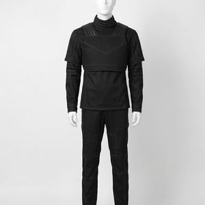 Pode incluir: Roupa t&aacute;tica preta exibida em um manequim. O conjunto inclui uma camisa de gola alta, um colete com detalhes em couro e cal&ccedil;as combinando. As costuras e os bolsos s&atilde;o detalhados com costuras brancas. A roupa &eacute; projetada para uma est&eacute;tica elegante e moderna.