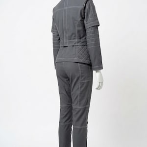 May include: A gray two-piece outfit on a mannequin. The jacket has white stitching and quilted detailing on the back. The pants have pockets and white stitching. The outfit is displayed on a black stand.