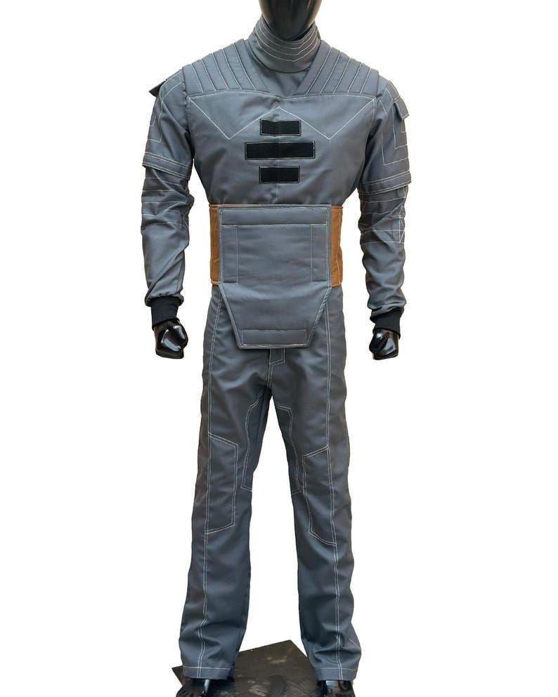 May include: A gray jumpsuit with white stitching, featuring a high collar and padded details on the shoulders and chest. The suit includes a brown belt and black gloves. The outfit is displayed on a mannequin.