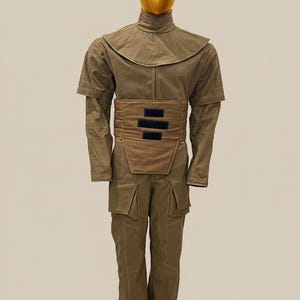 Puede incluir: Un disfraz de cuerpo entero en verde oliva, con cuello alto, capa en los hombros y cintur&oacute;n ancho con detalles negros. El atuendo incluye mangas largas con detalles estampados y pantalones a juego con bolsillos. Una cabeza de maniqu&iacute; dorada completa el conjunto.