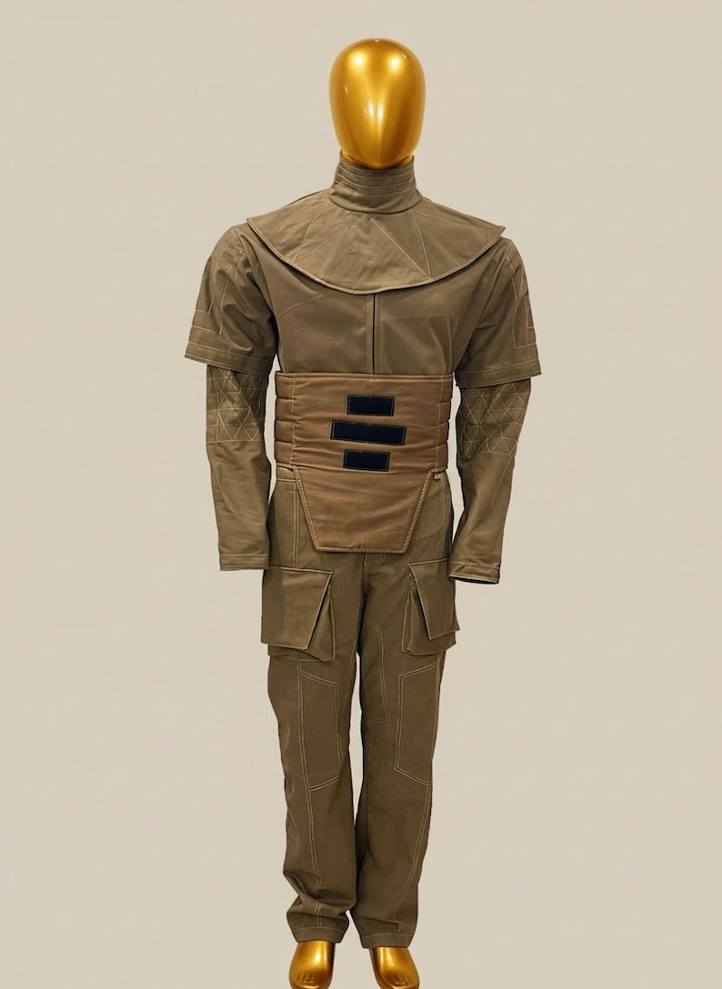 Puede incluir: Un disfraz de cuerpo entero en verde oliva, con cuello alto, capa en los hombros y cintur&oacute;n ancho con detalles negros. El atuendo incluye mangas largas con detalles estampados y pantalones a juego con bolsillos. Una cabeza de maniqu&iacute; dorada completa el conjunto.