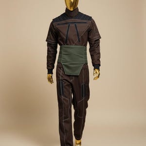 Puede incluir: Mono marr&oacute;n con detalles negros y costuras blancas, usado por un maniqu&iacute; con cabeza y manos doradas. El atuendo incluye un cuello alto, una faja verde y pantalones a juego. El maniqu&iacute; lleva zapatos dorados.