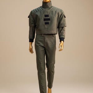 Puede incluir: Un conjunto verde de dos piezas sobre un maniqu&iacute;. La parte superior es una chaqueta corta con cuello alto y detalles negros. Los pantalones son de corte recto con costuras a juego. El maniqu&iacute; es de color dorado.