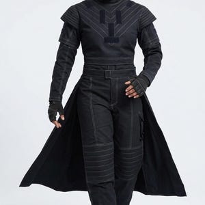 Puede incluir: Mono negro con costuras blancas. El atuendo presenta un cuello alto, detalles en los hombros y un elemento largo tipo falda. Los pantalones tienen rodilleras y bolsillos cargo. Guantes sin dedos completan el look.