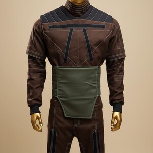 Puede incluir: Mono marr&oacute;n con detalles negros y costuras blancas, exhibido en un maniqu&iacute; con cabeza dorada. El atuendo presenta un cuello alto, hombros acolchados y un cintur&oacute;n verde. Los pantalones tienen paneles negros.