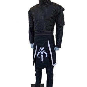 May include: Black costume with white accents, displayed on a mannequin. The outfit includes a high-collared jacket, pants, and a skirt-like piece with a white skull emblem. The jacket has vertical stitching and the pants have horizontal stitching.