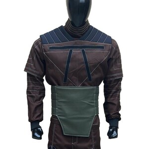 Puede incluir: Un disfraz marr&oacute;n con cuello alto, detalles en los hombros azul oscuro y ribetes negros. El disfraz presenta un cintur&oacute;n verde y pu&ntilde;os negros. El atuendo se exhibe en un maniqu&iacute;.