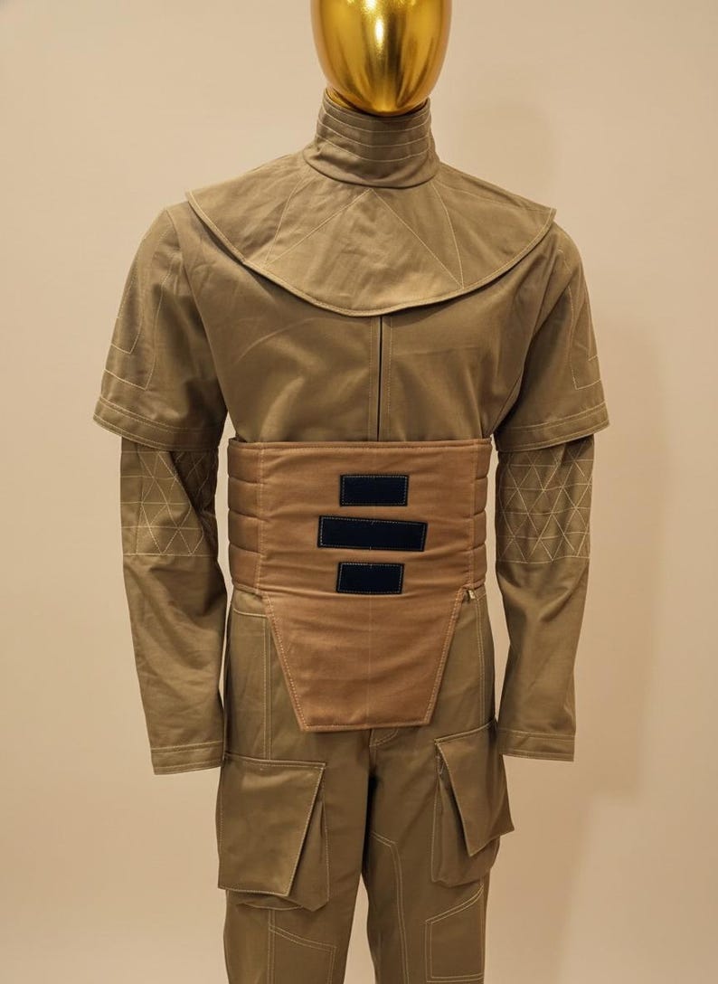 Puede incluir: Un disfraz de color marr&oacute;n con cuello alto y cintur&oacute;n ancho con tres rect&aacute;ngulos negros. El atuendo incluye mangas largas con costuras geom&eacute;tricas y pantalones a juego con bolsillos grandes. Un tocado dorado completa el conjunto.