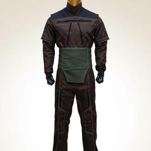 Puede incluir: Mono marr&oacute;n con detalles negros y una faja verde, exhibido en un maniqu&iacute; con cabeza dorada. El atuendo tiene un cuello alto y costuras visibles. Los pantalones tienen bolsillos.