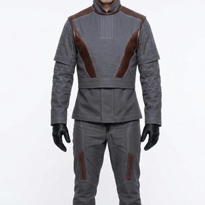 May include: A gray outfit with brown leather accents. The jacket has a high collar and quilted sleeves. The pants have brown leather patches on the knees. The person is wearing black gloves. The outfit is a futuristic design.