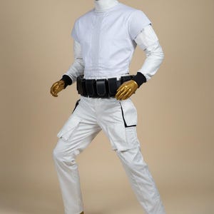 May include: A white futuristic costume featuring a high collar, short sleeves, and cargo trousers. The outfit includes a black belt with pouches and black wrist cuffs. The mannequin's head, hands, and feet are gold.
