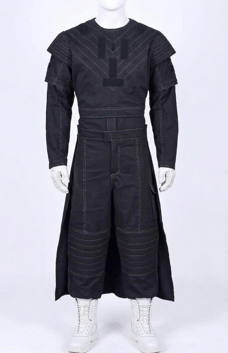 Puede incluir: Traje negro con costuras blancas, que incluye una parte superior de manga larga con detalles en los hombros y cintura con cintur&oacute;n, adem&aacute;s de pantalones con bolsillos de carga y acolchado en las rodillas. Botas blancas completan el conjunto. El traje tiene un dise&ntilde;o futurista.