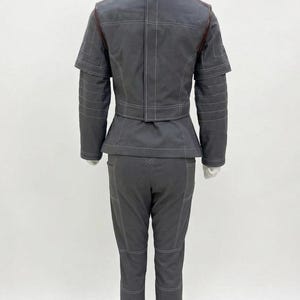 May include: A gray two-piece outfit on a mannequin. The jacket has a high collar and a peplum waist, with white stitching and brown accents. The matching pants have white stitching and knee patches. The outfit is displayed on a white background.