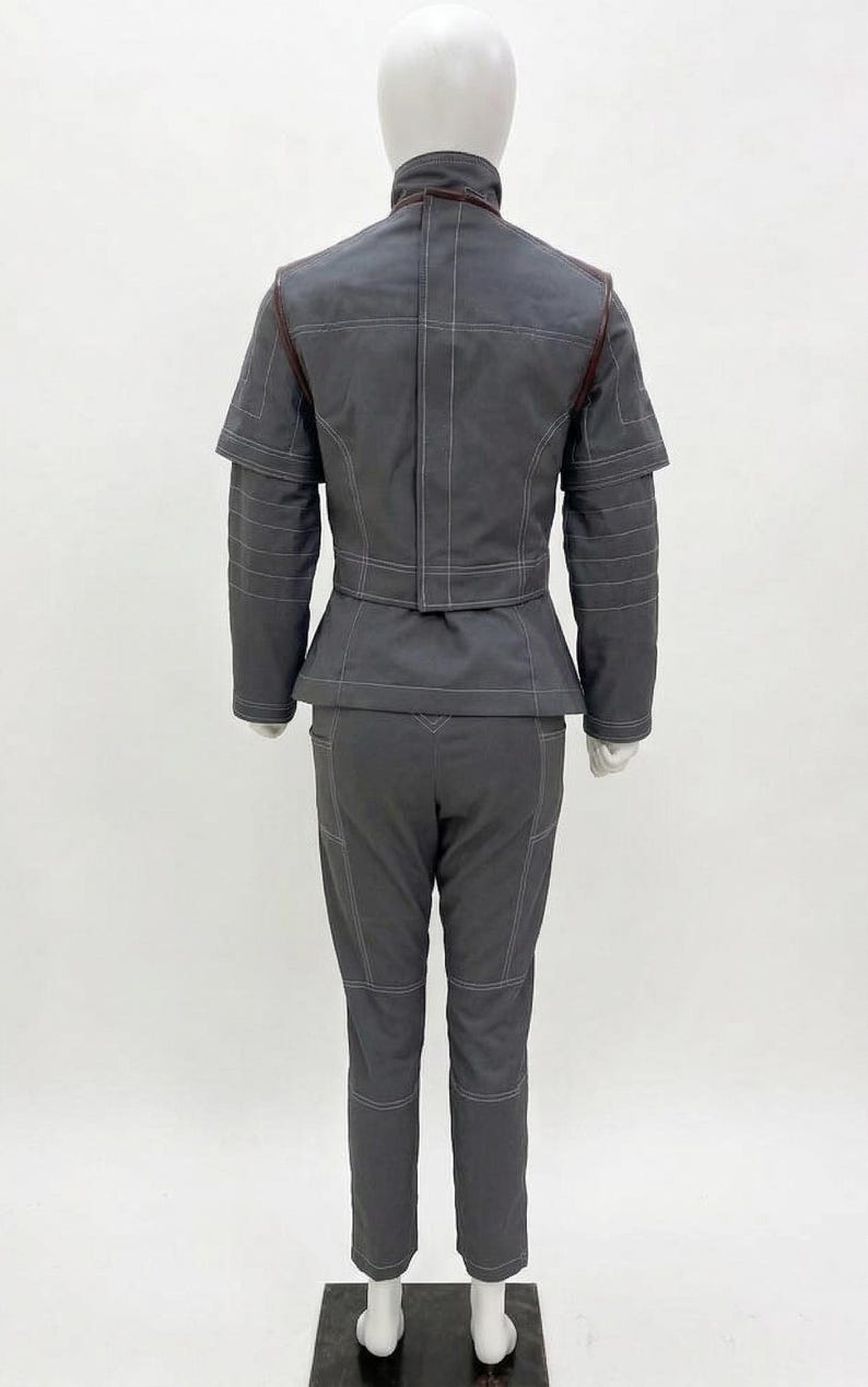 May include: A gray two-piece outfit on a mannequin. The jacket has a high collar and a peplum waist, with white stitching and brown accents. The matching pants have white stitching and knee patches. The outfit is displayed on a white background.
