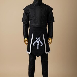 May include: Black costume with white accents, displayed on a mannequin. The outfit includes a high-collared top, short-sleeved jacket, and pants. A black apron with a white emblem is also part of the ensemble. The mannequin has a gold head and hands.