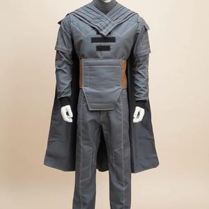 May include: A gray costume displayed on a mannequin. The outfit includes a long-sleeved jacket with a high collar, a cape, pants, and a brown belt. White stitching details the seams. The costume is likely for cosplay or a theatrical production.