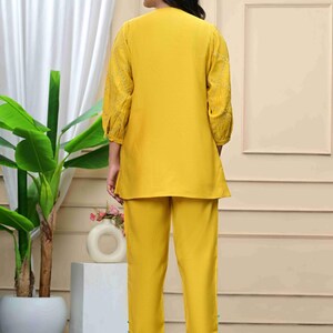 May include: A two-piece yellow outfit, consisting of a tunic top and matching pants. The tunic has three-quarter sleeves with embroidered detailing. The pants are straight-legged. The outfit is worn with clear heeled sandals. The background includes a plant and a white curtain.