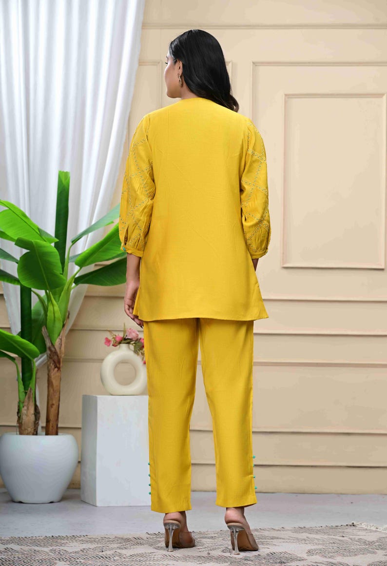 May include: A two-piece yellow outfit, consisting of a tunic top and matching pants. The tunic has three-quarter sleeves with embroidered detailing. The pants are straight-legged. The outfit is worn with clear heeled sandals. The background includes a plant and a white curtain.