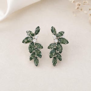 May include: A pair of silver-toned earrings featuring a cluster of marquise-cut moss green stones arranged in a leaf-like pattern, accented with small, clear, round-cut stones set within the metal frame to create a botanical-inspired design.