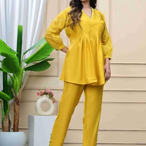 May include: A two-piece yellow outfit featuring a tunic top and matching pants. The top has a V-neck, pleated front, and embroidered sleeves. The pants have a straight leg design with button details at the ankles. The outfit is made of a soft, flowing fabric.
