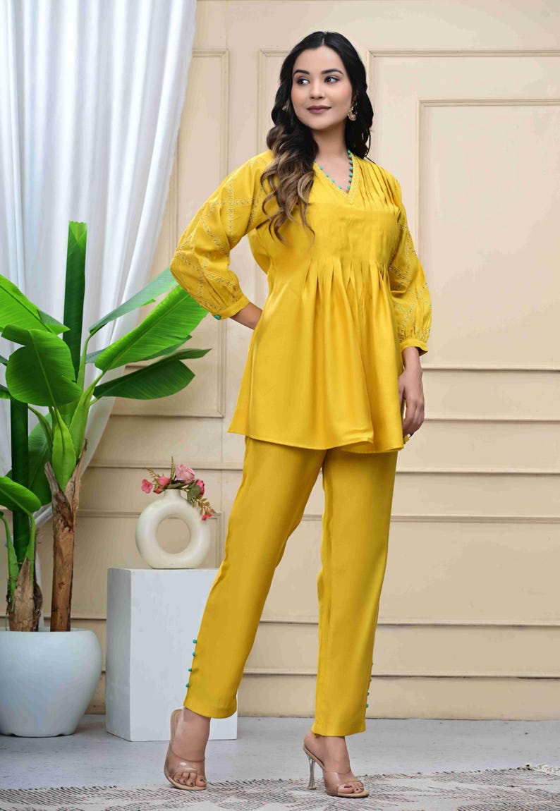 May include: A two-piece yellow outfit featuring a tunic top and matching pants. The top has a V-neck, pleated front, and embroidered sleeves. The pants have a straight leg design with button details at the ankles. The outfit is made of a soft, flowing fabric.