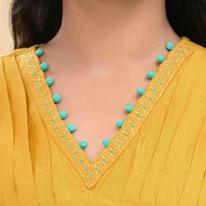 May include: Mustard yellow top with a V-neckline. The neckline is adorned with turquoise pom-poms and embroidered detailing. The fabric has vertical pleats, adding texture to the garment. The top is a close-up shot, focusing on the neckline and upper chest area.