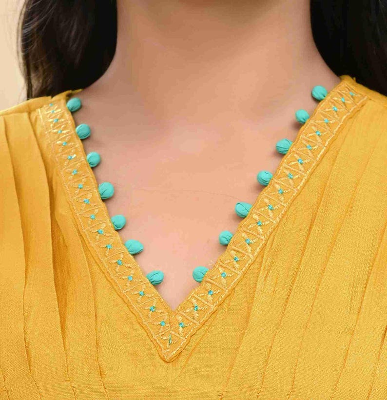 May include: Mustard yellow top with a V-neckline. The neckline is adorned with turquoise pom-poms and embroidered detailing. The fabric has vertical pleats, adding texture to the garment. The top is a close-up shot, focusing on the neckline and upper chest area.