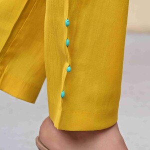 May include: Close-up of yellow pants with turquoise buttons. The pants are made of a smooth fabric and have a tailored fit. The buttons are small and round, adding a pop of color to the design.