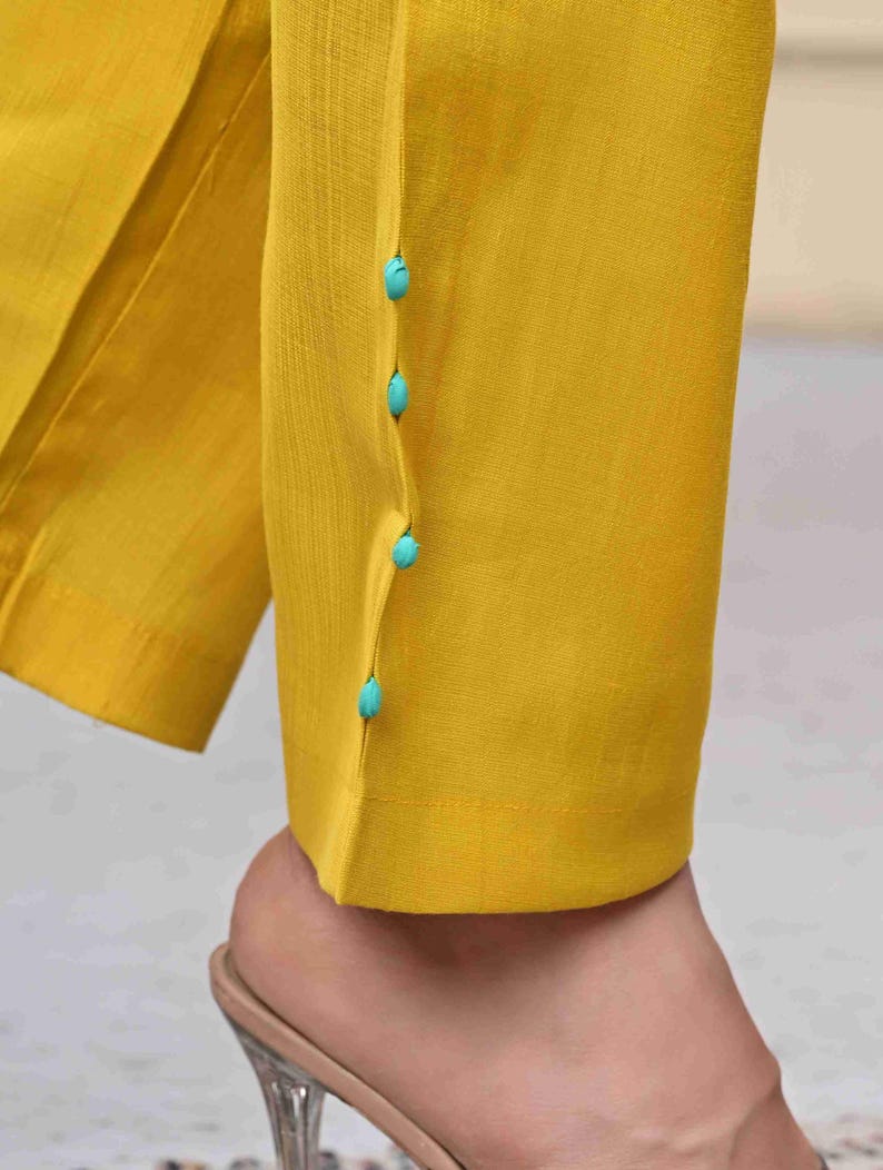 May include: Close-up of yellow pants with turquoise buttons. The pants are made of a smooth fabric and have a tailored fit. The buttons are small and round, adding a pop of color to the design.