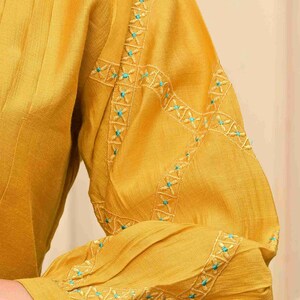 May include: Mustard yellow long-sleeved top with intricate turquoise embroidery on the sleeves. The sleeve cuff features a turquoise button. The fabric has a subtle texture, and the design is elegant.