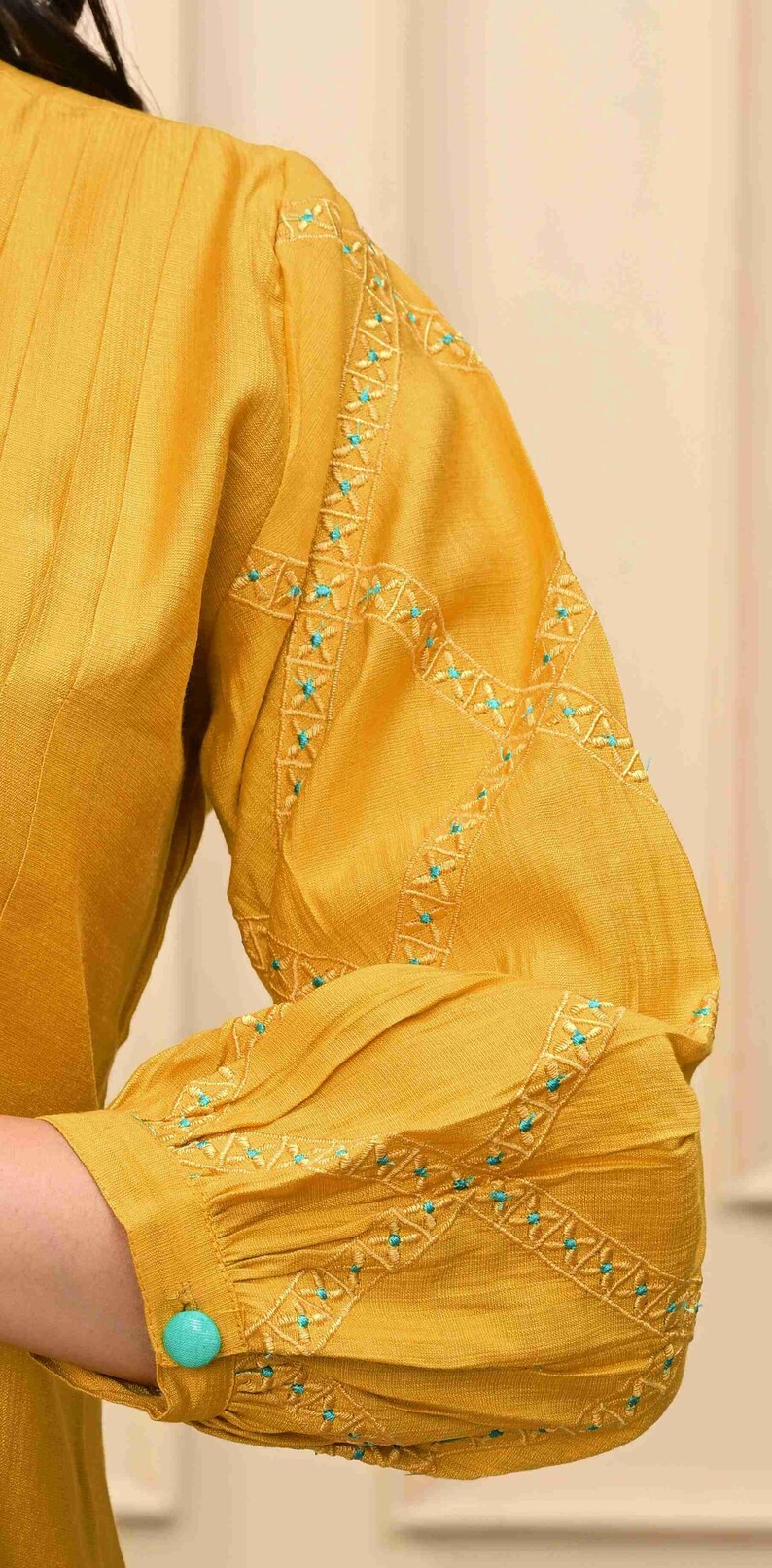 May include: Mustard yellow long-sleeved top with intricate turquoise embroidery on the sleeves. The sleeve cuff features a turquoise button. The fabric has a subtle texture, and the design is elegant.