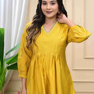 May include: Mustard yellow tunic and pants set. The tunic features a V-neckline with turquoise bead accents, pleats, and embroidered sleeves. The outfit is made of a soft, flowing fabric.