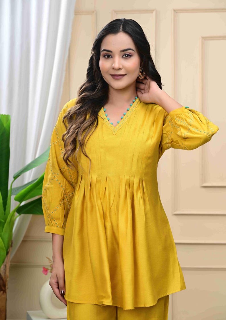 May include: Mustard yellow tunic and pants set. The tunic features a V-neckline with turquoise bead accents, pleats, and embroidered sleeves. The outfit is made of a soft, flowing fabric.