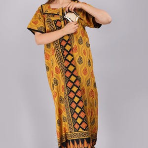 Könnte beinhalten: Ein bodenlanges Kaftan-Kleid in Senfgelb mit einem schwarzen und roten geometrischen Rautenmuster in der Mitte und einem durchgehenden schwarzen und roten Blattdruck. Das Kleidungsstück hat kurze Ärmel mit schwarzem Saum und einen eckigen Ausschnitt.